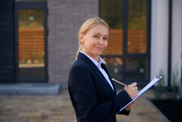 Cheerful businesswoman standing near office and writing capital improved value on paper attached to clipboard which she holding in hands