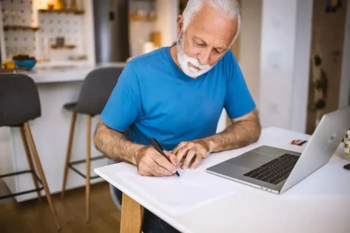 Commercial property landlord in blue shirt at home table, making list of on site inspection items for his commercial property investment. The mature man with white short beard has a laptop open in front of him.