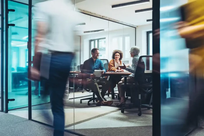 Couple having a meeting in modern boardroom with professional woman with short grey hair, while colleagues walk past in corridor.