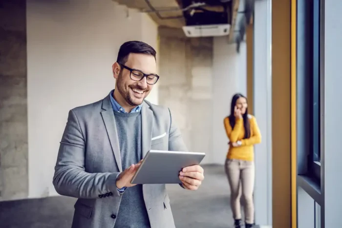 Realtor smiling in front of his tablet while a client speaks on the phone in the background.